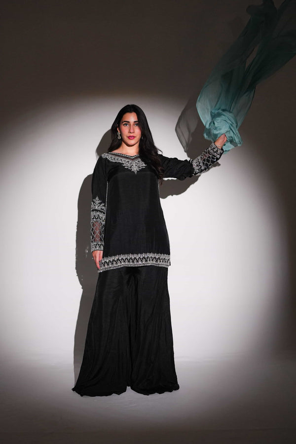 Woman in a black traditional outfit with intricate patterns holding a blue fabric against a gray background